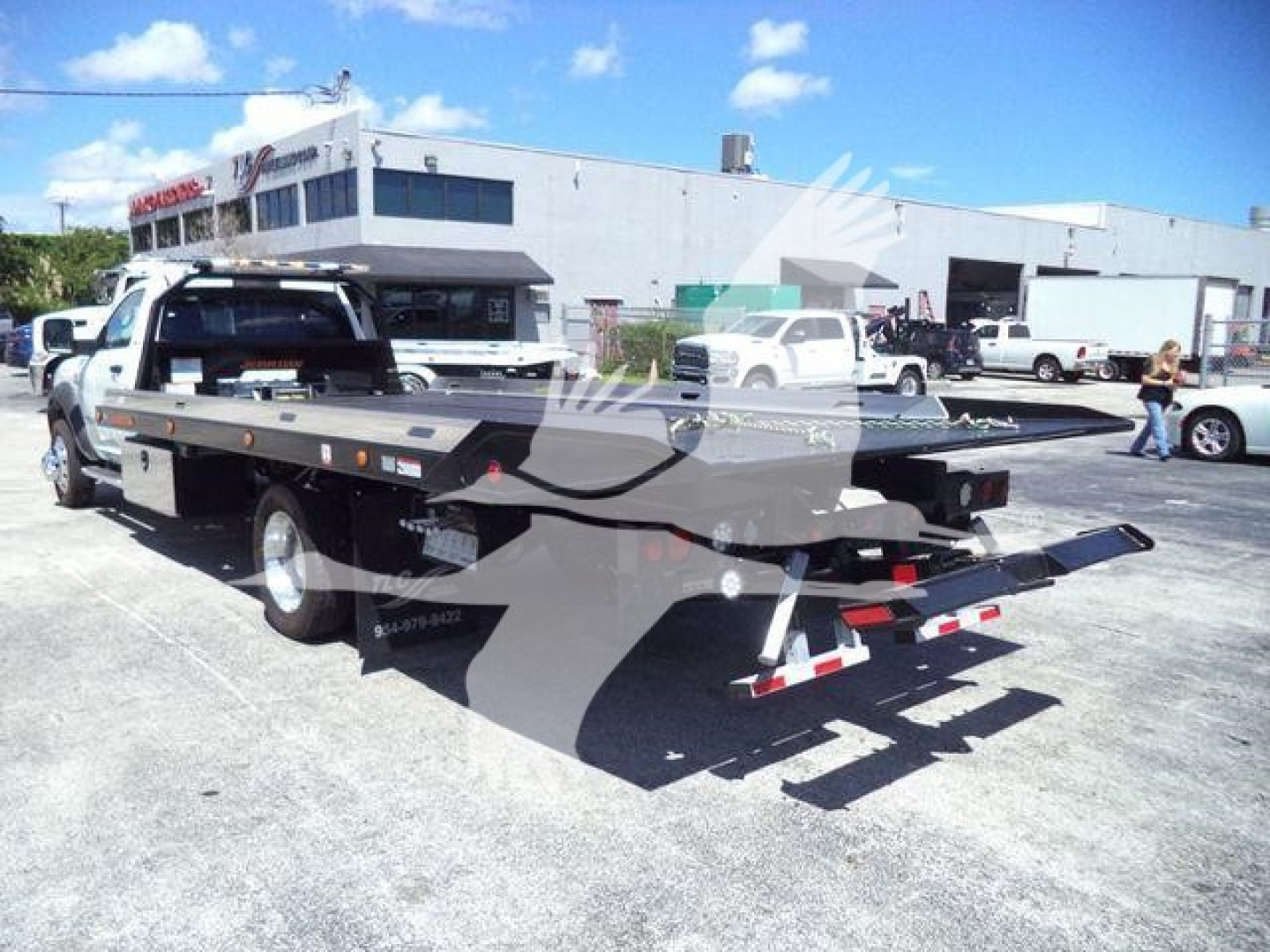 Flatbed rollback tow truck on dealership lot, rear view shows ramp and winch, Ram 5500 SLT hauling bed with dual rear wheels and equipment toward back.