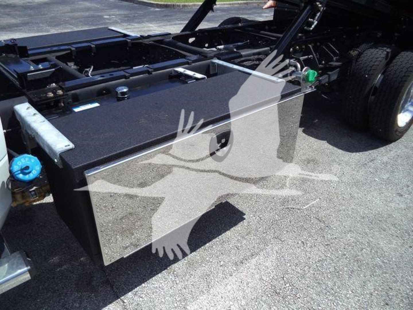 Truck chassis with a large rectangular metal toolbox mounted near the cab; visible components include a blue reservoir cap, battery area, and wheel on the right; no legible branding text is clearly readable on the toolbox.