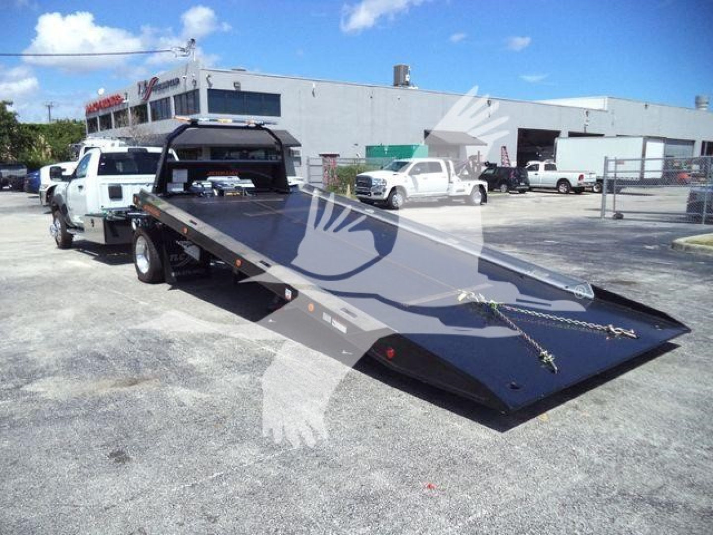 Ram 5500 rollback tow truck with a heavy steel flatbed, white pickup in background, dealership lot, no visible text on the bed.