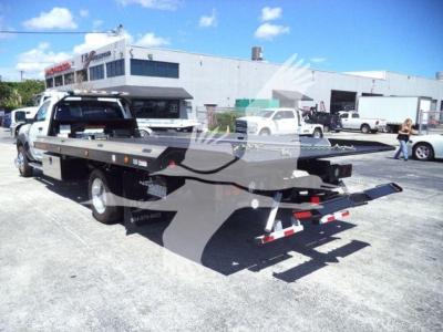 Flatbed rollback tow truck on dealership lot, rear view shows ramp and winch, Ram 5500 SLT hauling bed with dual rear wheels and equipment toward back.