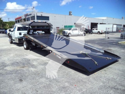 Ram 5500 rollback tow truck with a heavy steel flatbed, white pickup in background, dealership lot, no visible text on the bed.
