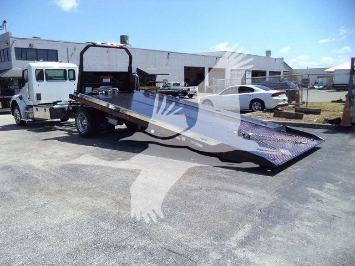 Flatbed rollback truck with a long lowered ramp in a yard; cab is white; equipment has Kaw hillside logo overlay; background shows other vehicles and a low industrial building.
