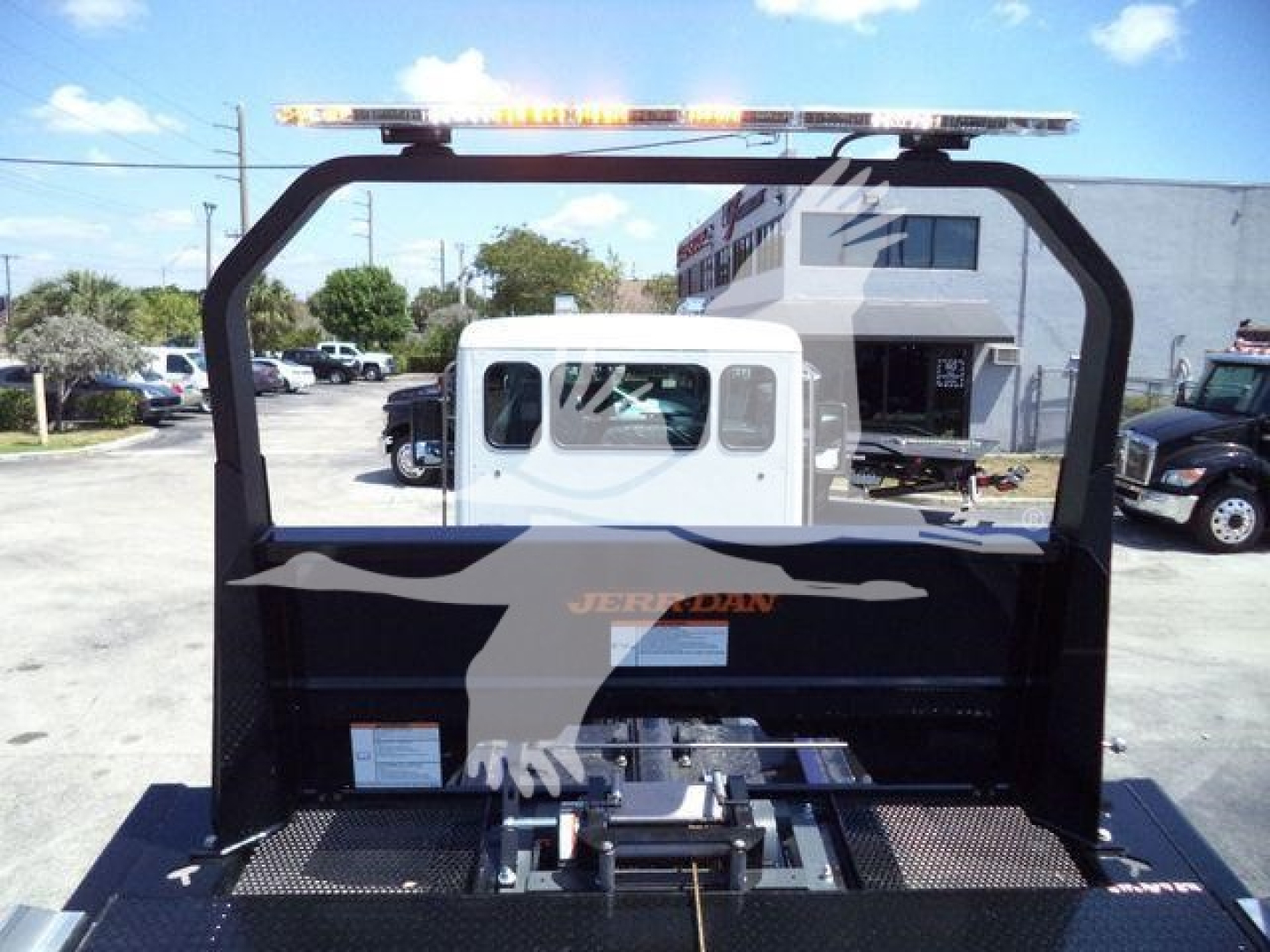 Truck bed assembly for a rollback truck with a tall frame and orange warning lights on top; rear cab window visible; mounting equipment in bed area.
