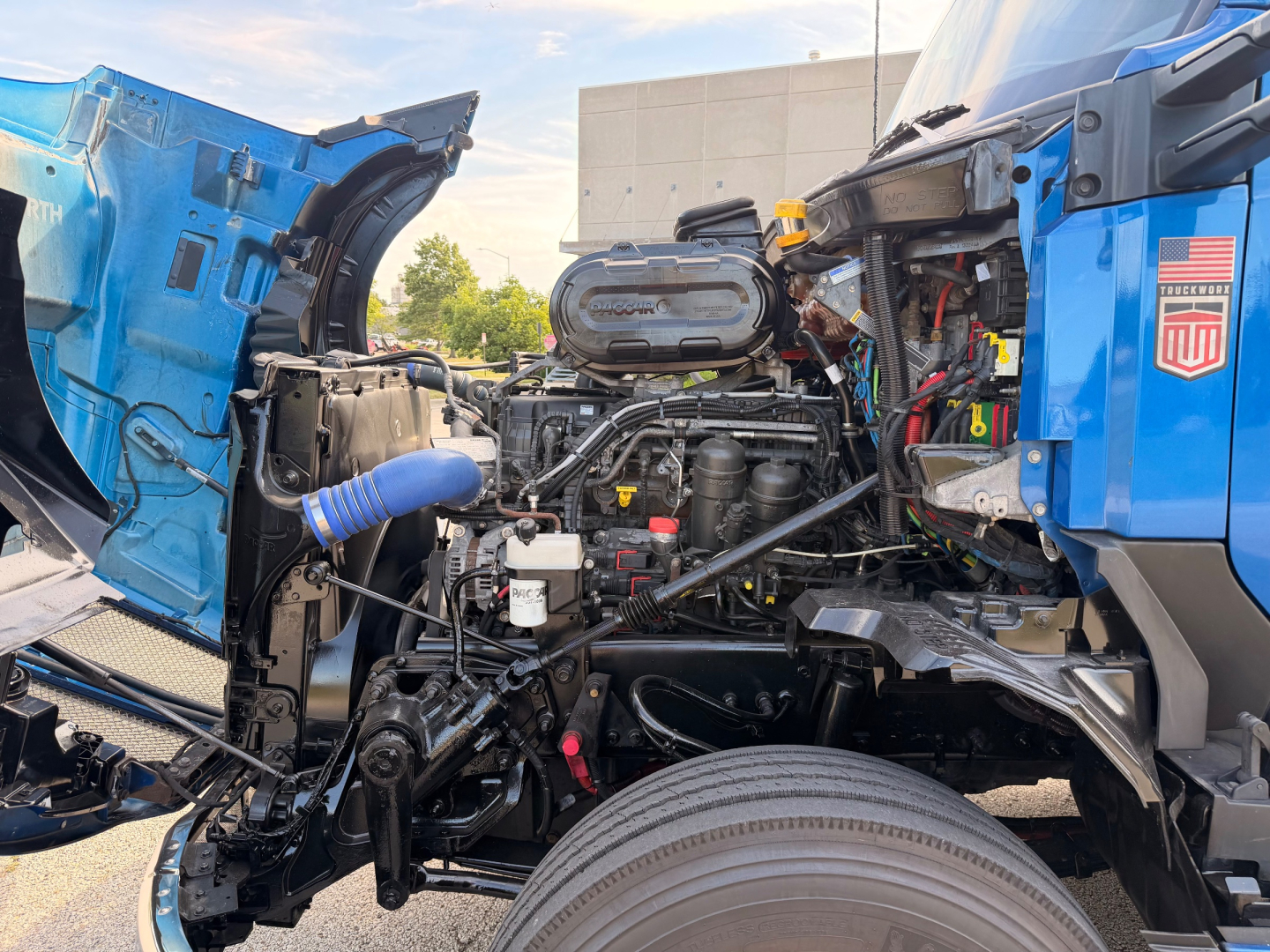 Truck engine bay of a blue 2022 Kenworth T680 with hood open, showing engine, hoses, and components.