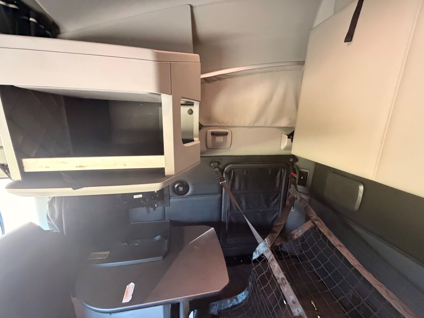 Interior cab view of a 2022 Kenworth T680 sleeper with a microwave, small table, and beige upholstery; visible seat belts and storage net.