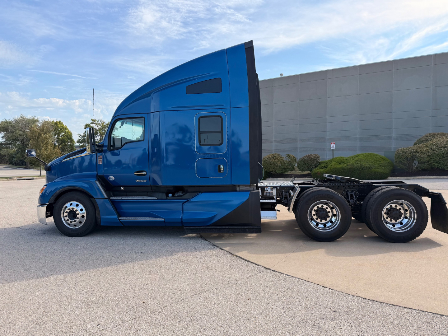 Kenworth T680 tractor unit side view in blue, cab with sleeper, dual rear axles, empty trailer chassis, factory wheels visible.