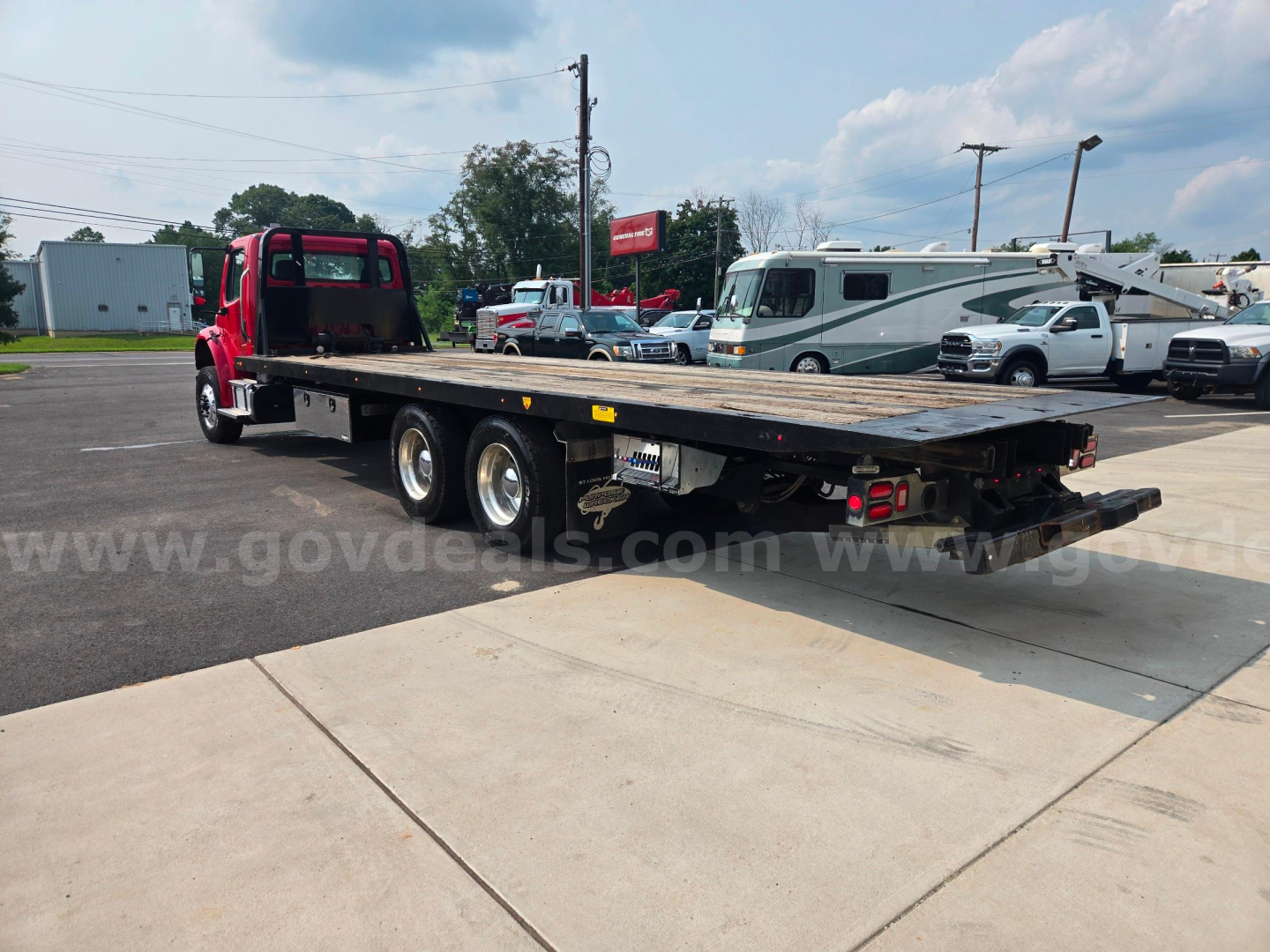 Flatbed tow truck from rear left side showing long wood deck with triple axle and red cab, Freightliner M2 platform truck.
