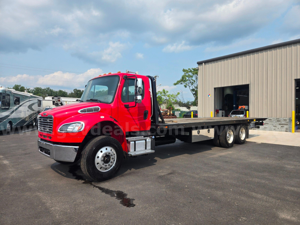 Flatbed tow truck in red, Freightliner M2 chassis, idle in lot with service shop in background.