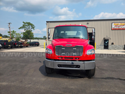 Front view of a red 2024 Freightliner M2 tow truck in a parking lot, showing large grille, headlights, bumper, and side mirrors.