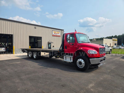 Red Freightliner M2 tow truck with flatbed in front of a beige industrial building; stock number signage and clear blue sky.