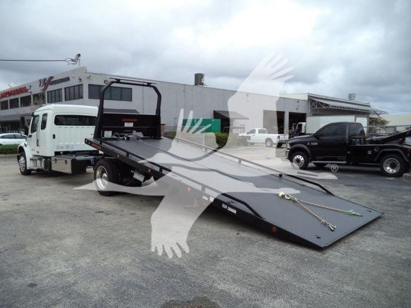 Truck with a flatbed ramp in a dealership lot showing Freightliner M2 106 rollback bed and white cab