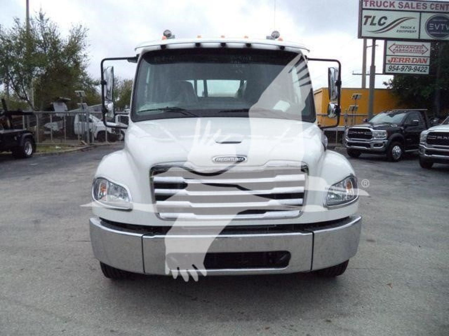 Front view of a white Freightliner M2 106 rollback truck showing grille, bumper, and windshield with dealership lot in background.