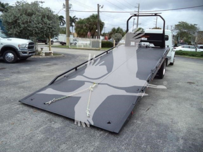 Flatbed tow truck with a lowered, steel ramp bed and chain tie-downs, viewed from the front left, light gray surface, Freightliner M2 106 in background.