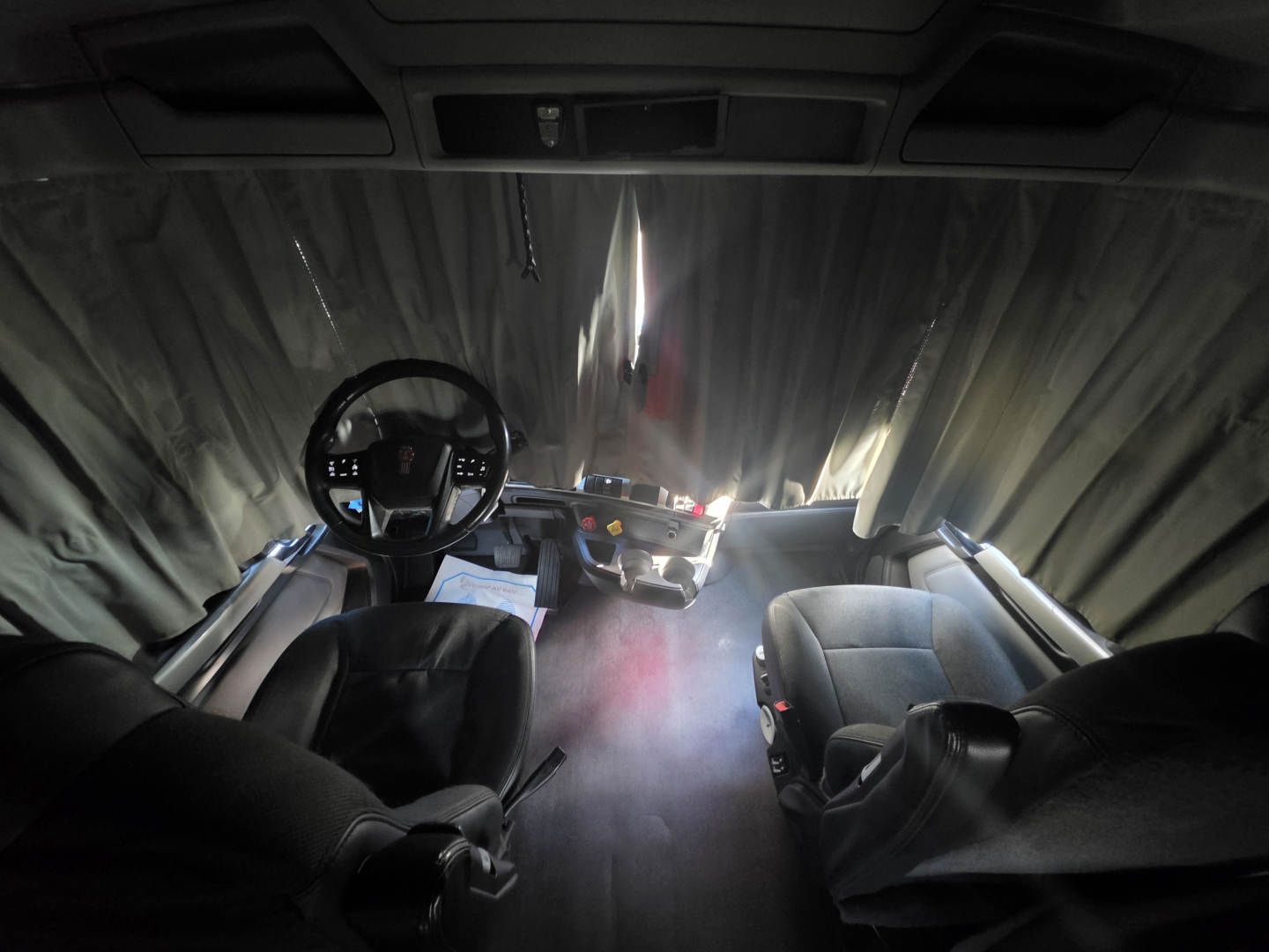 Cab interior of a 2023 Kenworth T680 truck showing driver and passenger seats, steering wheel, dashboard, and window divider curtain.