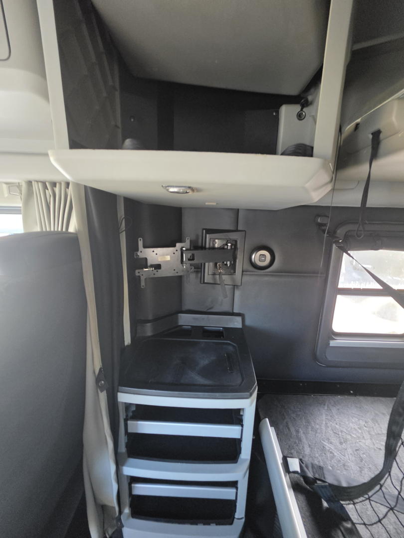 Kenworth T680 sleeper cab interior showing a mounted CB/monitor bracket and storage drawers beneath a small desk area, cab side window present.