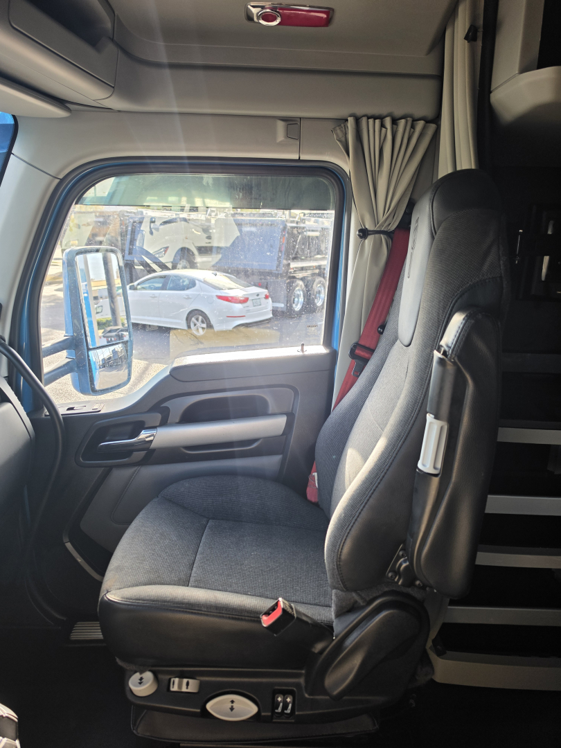 Truck cab interior of a Kenworth T680 with adjustable seat, door panel, window, and curtains; view through the driver window shows other trucks in a lot.