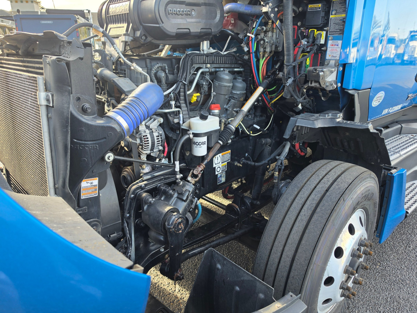 Kenworth T680 engine bay showing turbo, hoses, coolant reservoir, and various hoses wires with blue truck body and front wheel.