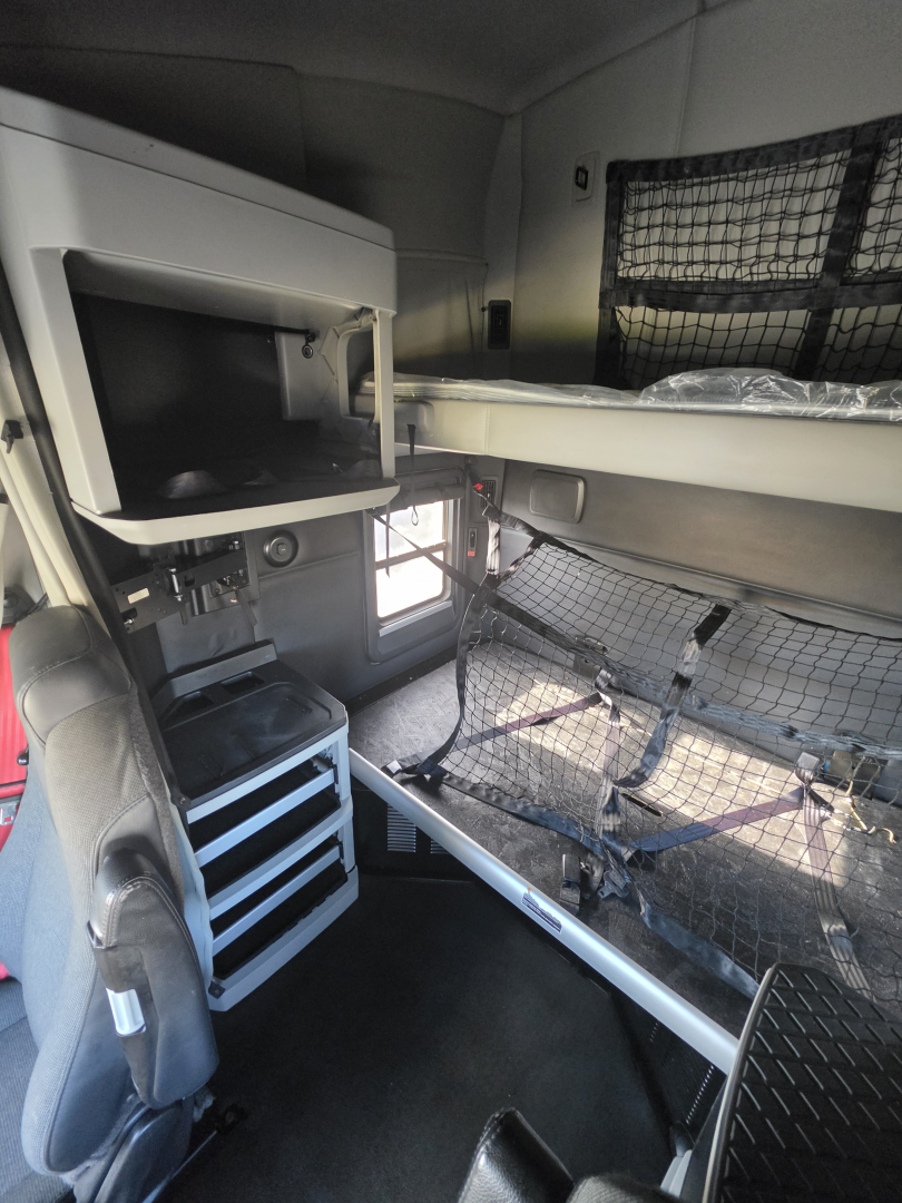 Cab interior of a 2023 Kenworth T680 sleeper with bunk area and overhead storage, driver seat, toolbox, and mesh partitions visible.