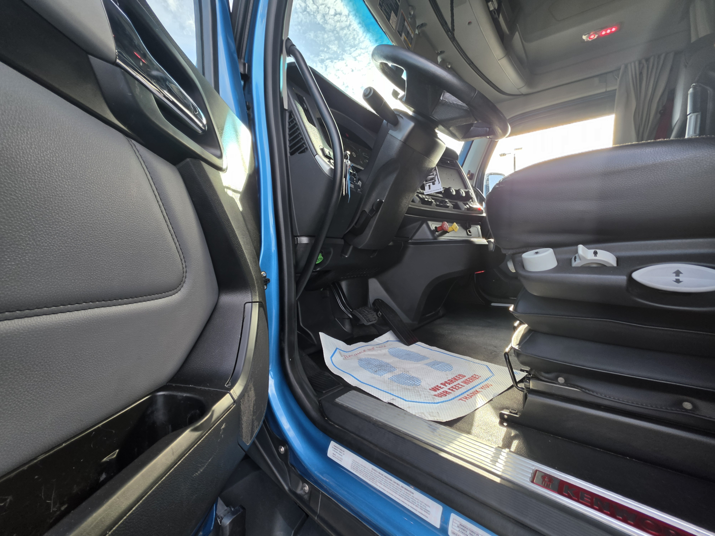 Kenworth T680 truck interior view from driver door with steering column, dashboard, and driver's seat. Visible are pedal area, floor mats, and entry step.