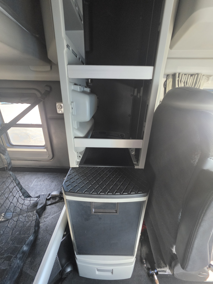 Cab interior of a 2023 Kenworth T680 with overhead storage rack, cabinet area, and steps; focus on a small cabinet with ladder-like rack, and a chair seat visible.