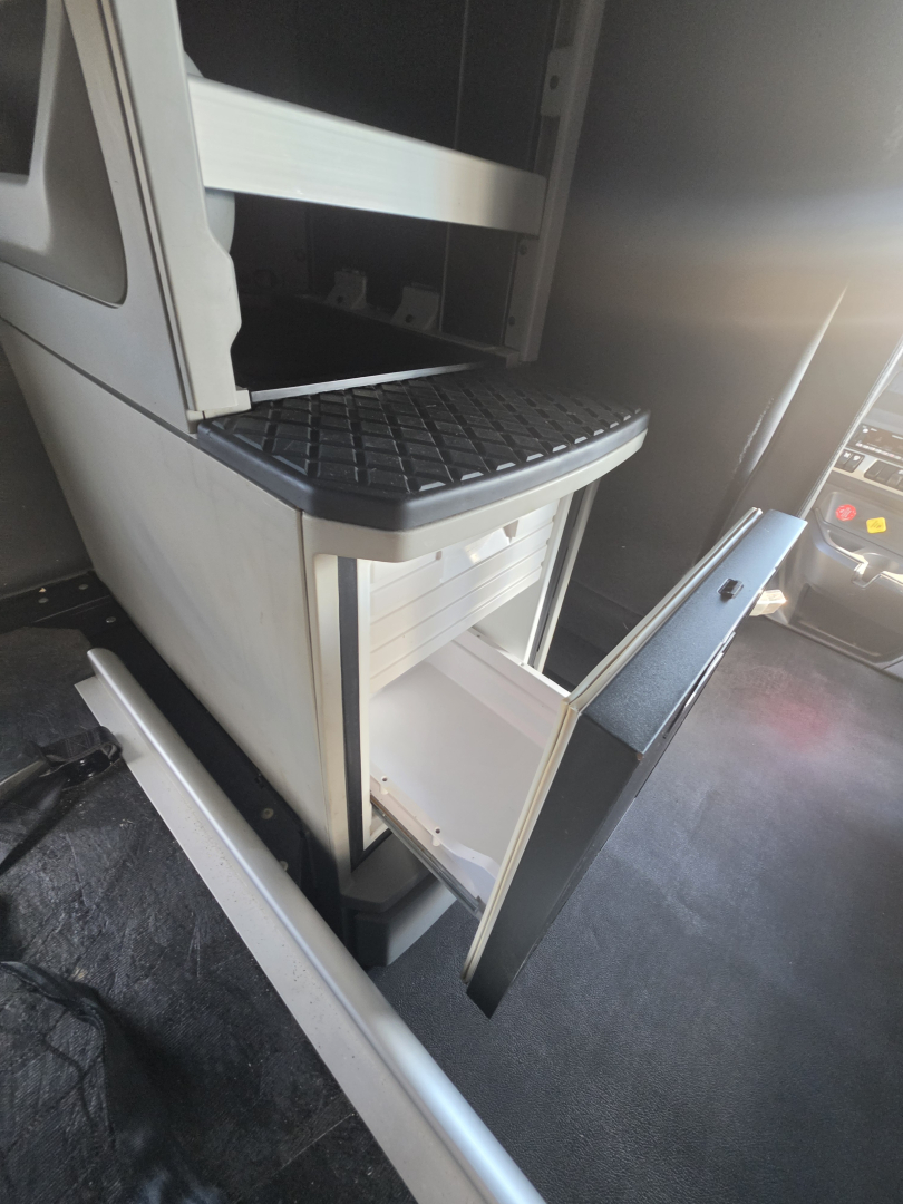 Truck interior showing a small open storage drawer under a step with a textured black tread plate; nearby is a control panel and door edge.