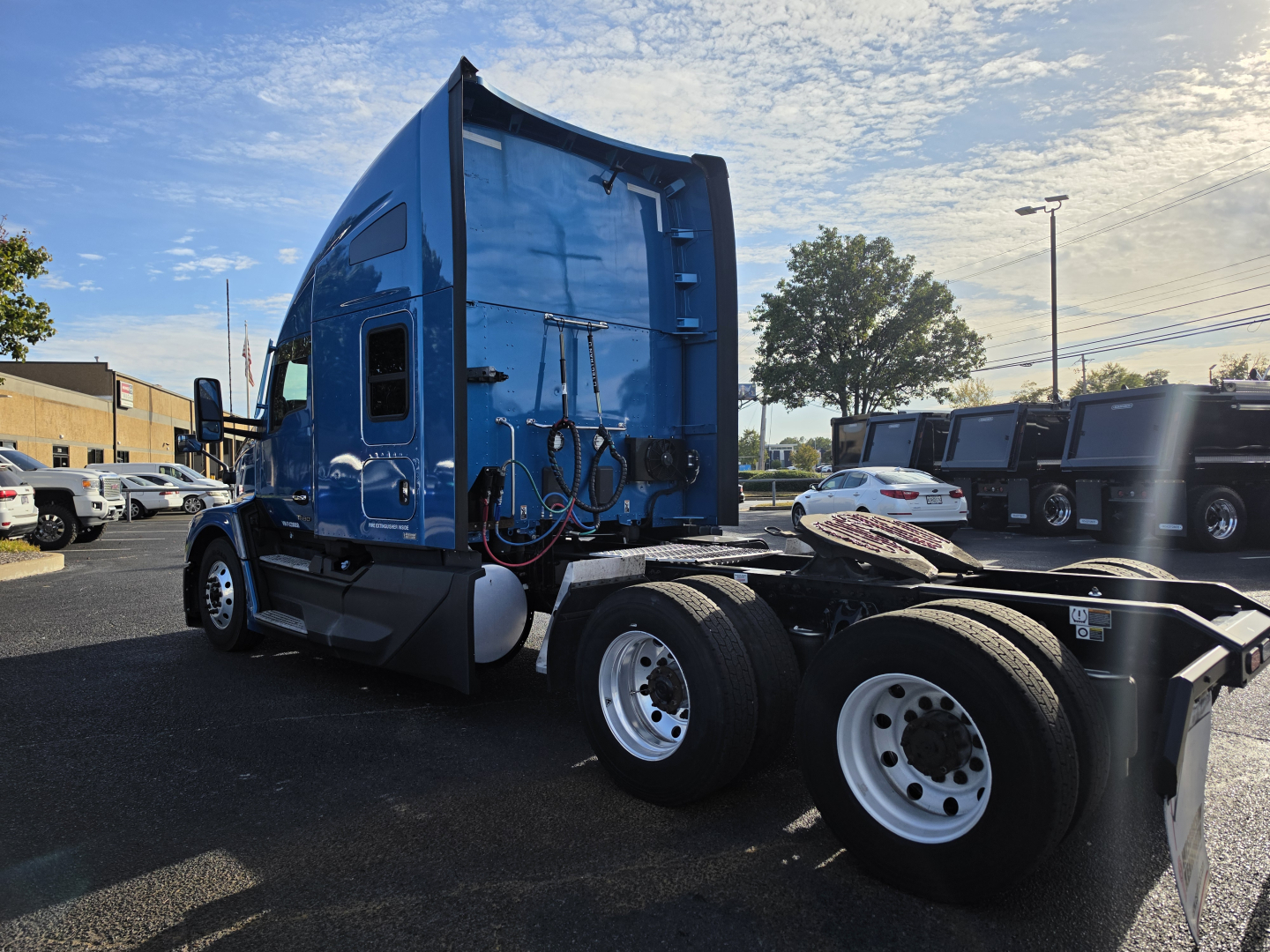 Kenworth T680 truck side view with blue cab and dual rear axles, visible fuel tanks and wiring; dealership lot in background.