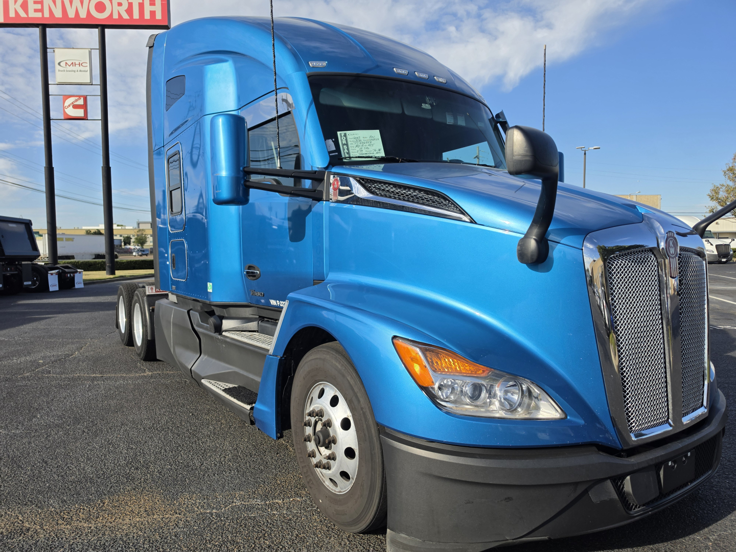 Blue 2023 Kenworth T680 conventional sleeper truck showing front grille and left-side cab, with Kenworth signage in background.