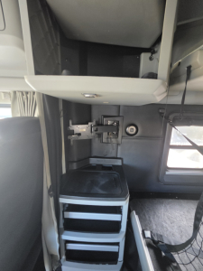 Kenworth T680 sleeper cab interior showing a mounted CB/monitor bracket and storage drawers beneath a small desk area, cab side window present.