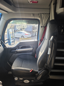 Truck cab interior of a Kenworth T680 with adjustable seat, door panel, window, and curtains; view through the driver window shows other trucks in a lot.