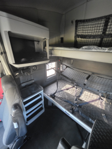Cab interior of a 2023 Kenworth T680 sleeper with bunk area and overhead storage, driver seat, toolbox, and mesh partitions visible.