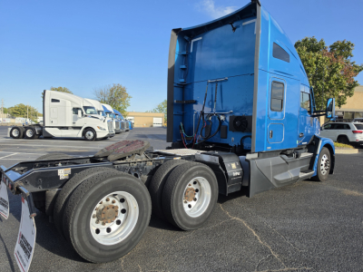 Kenworth T680 truck cab in blue, rear view of chassis showing dual rear axles and fifth wheel plate; other trucks in lot in background.