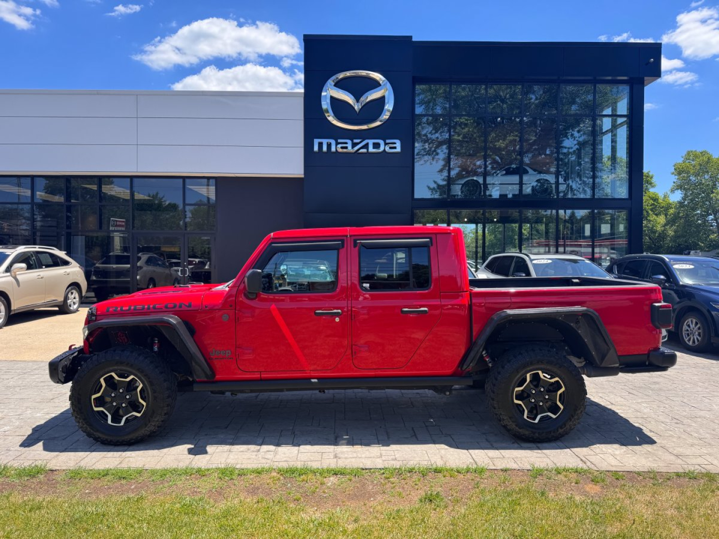 Red 2021 Jeep Gladiator pickup truck parked in front of Mazda dealership building with large glass windows and Mazda sign.