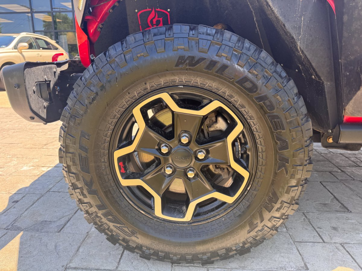 Jeep Gladiator wheel and tire close-up showing Nitto Mud Grappler tire and black gold-accented rim; part of front fender and brake components visible.