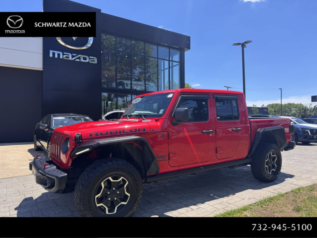Jeep Gladiator Rubicon crew cab pickup truck in red with black rims parked in dealership lot, Mazda building in background.