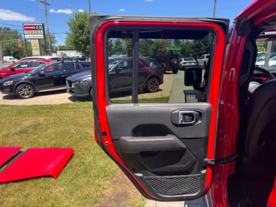 Jeep Gladiator rear passenger door panel open showing interior door handle, armrest, storage pocket; red exterior paint visible; car dealership lot in background.