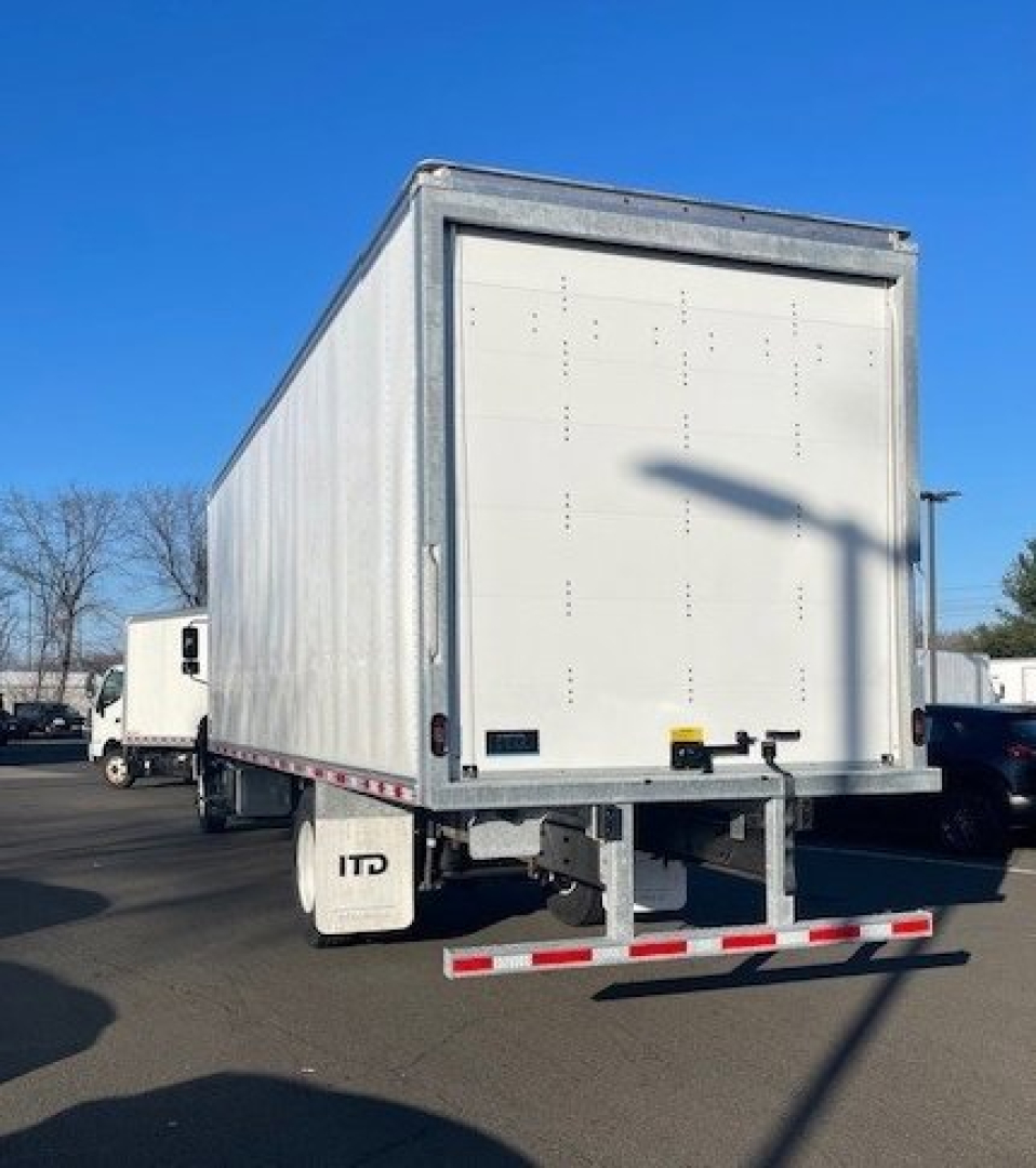 Box van truck rear view showing box body, rear doors, and dual rear tires; white cargo trailer with reflective tape and bumper; sunny day.