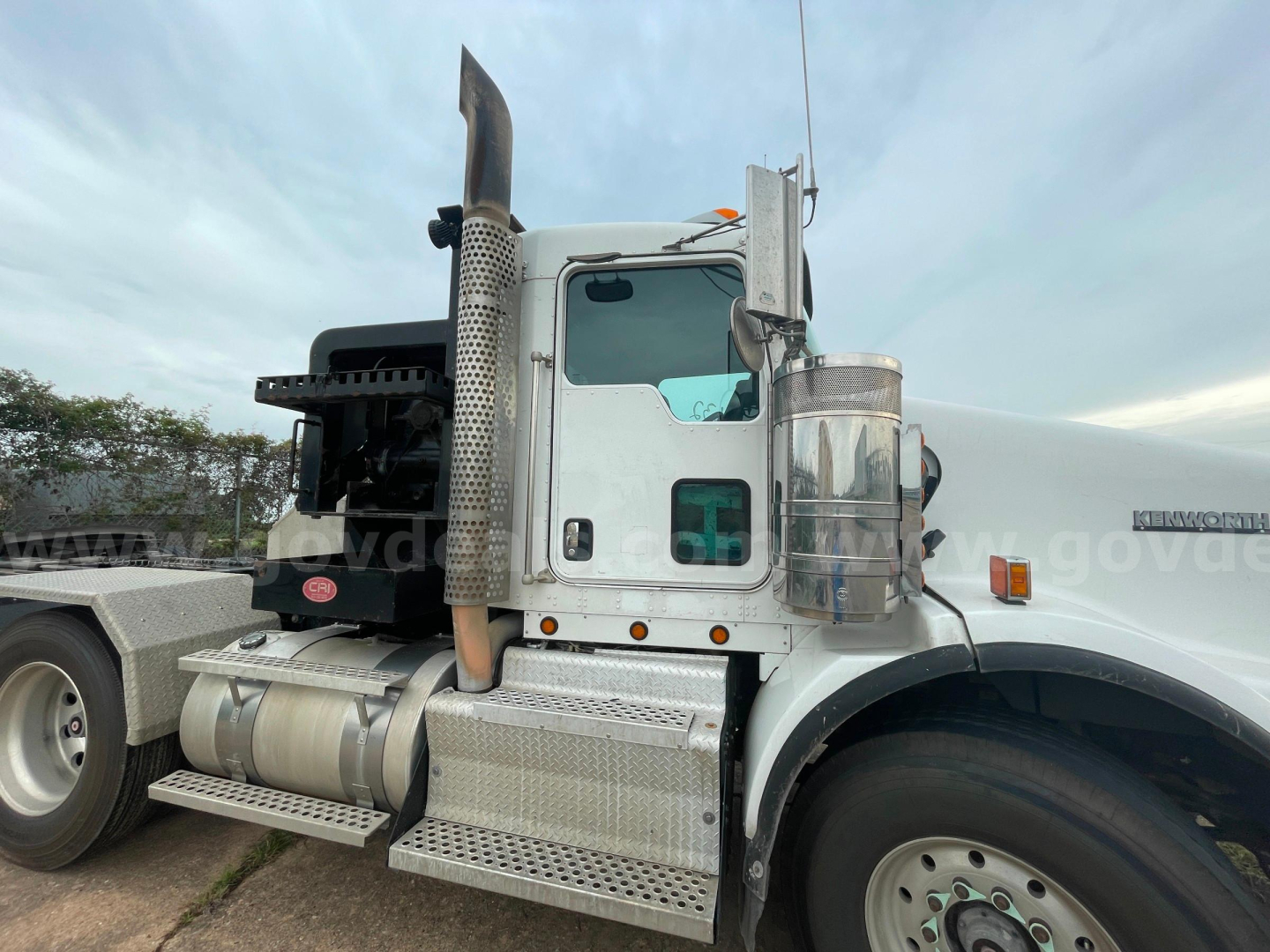Kenworth T800 cab view showing chrome fuel tanks, side step, and exhaust stack; front wheel and part of sleeper cabin visible.