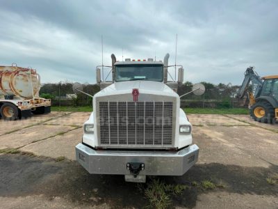 Kenworth T800 front view showing large chrome grille, bumper, and hood with Kenworth emblem; white cab and grey bumper; trucks in lot background.