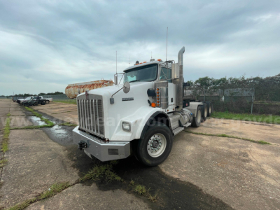 Kenworth T800 semi-truck in a lot, white cab, front grille with vertical bars, dual headlights, chrome exhaust stacks, dual rear axles, bumper, and orange marker lights.