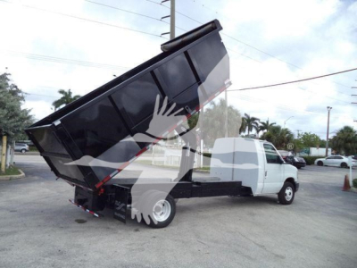 Ford E-450 dump truck with tilted hydraulic dump bed in a dealership lot. Visible text includes license plate-like sticker and VIN label on the windshield area? The image shows a white cab and black dump bed raised high.