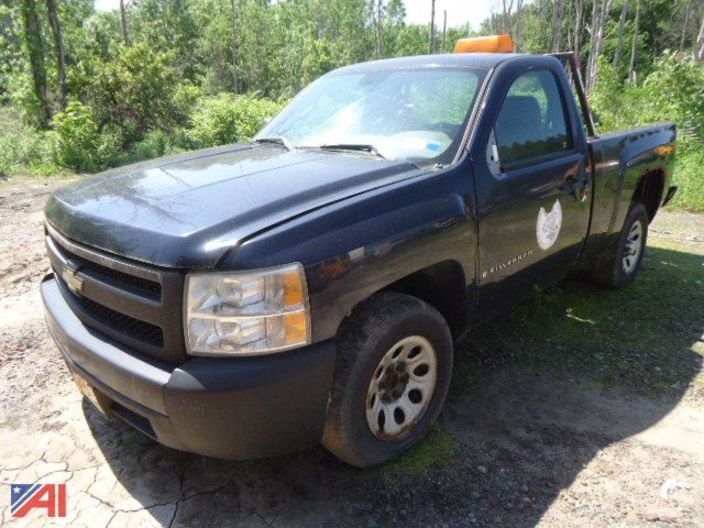 Chevrolet Silverado pickup truck in black/blue shade, single cab, cab with police-style emblem on door, orange beacon on roof, front view with highway dirt surrounding, daytime.