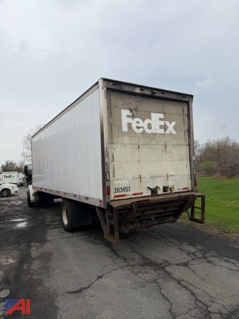 Box truck rear view showing FedEx-branded trailer with closed white cargo box and dark dock bumper. License/ID tag 203457 visible on rear. Overcast sky and road surface.