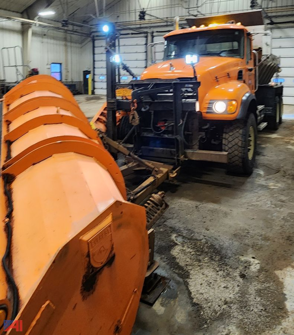 Orange Mack CV 713 dump truck with plow and sander inside a workshop; front grill, headlights, and large snowplow blades visible.
