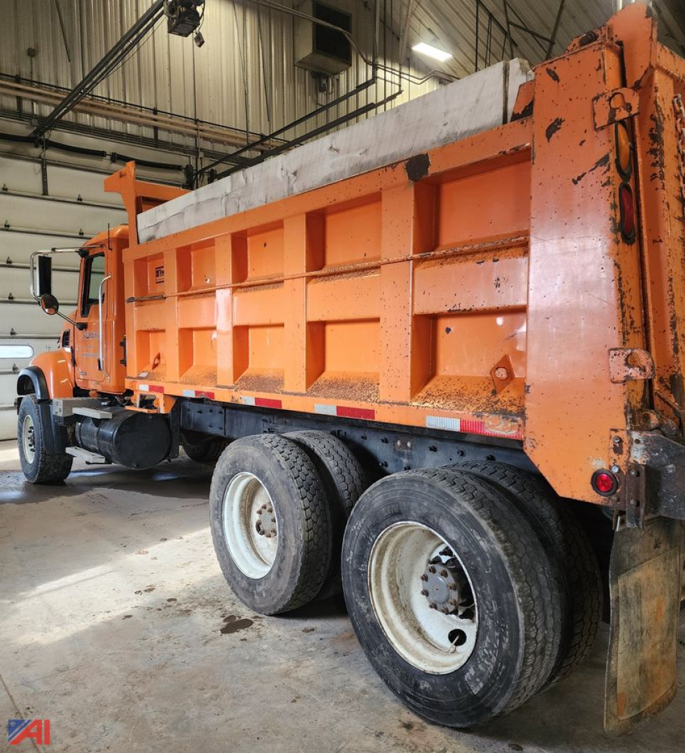 Dump truck side view showing orange Mack CV 713 with multiple rear axles and plow/sander, inside a warehouse.