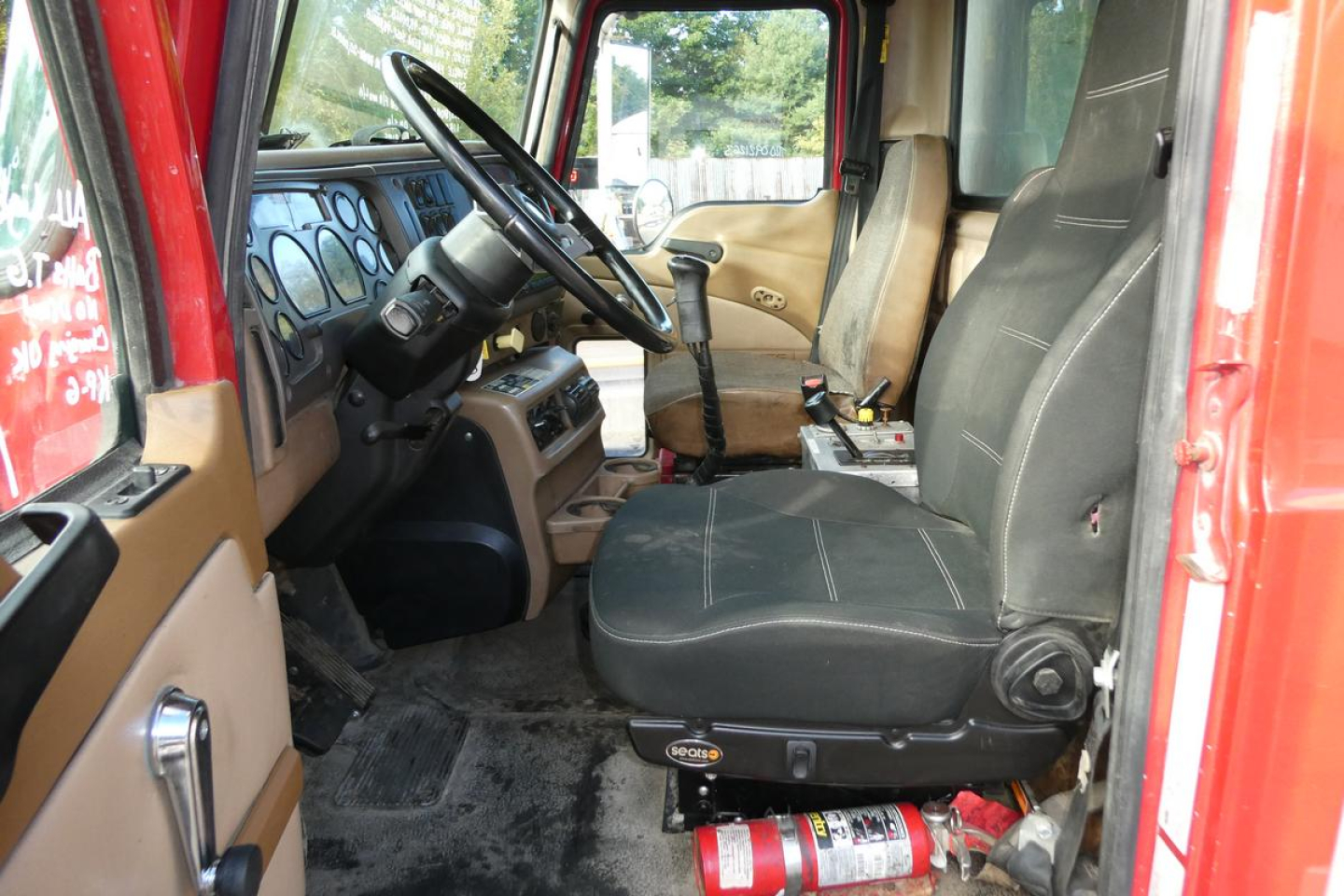 CV truck cab interior showing driver seat and steering wheel with beige door panels and dashboard; fire extinguisher under seat; gear shifter and pedals visible.