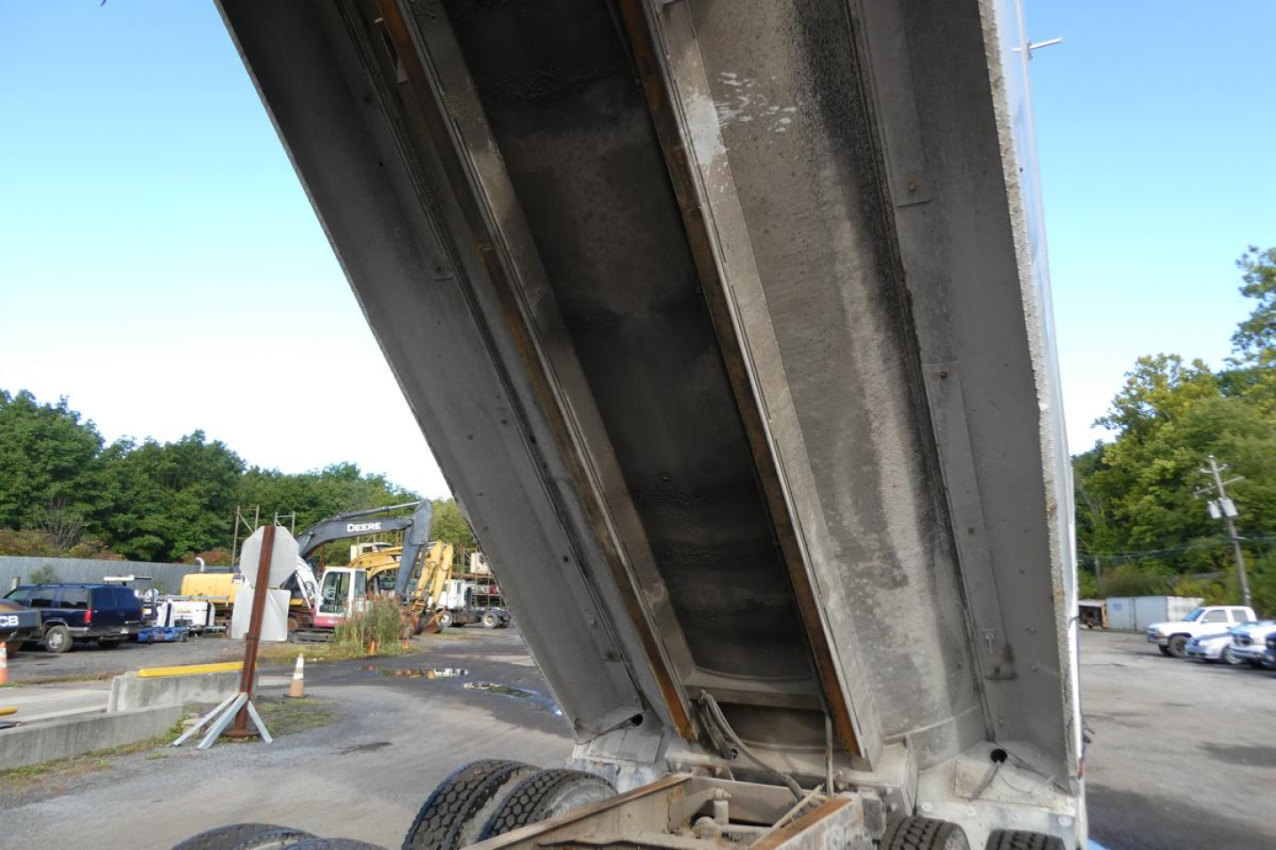 Dump truck bed raised showing metal inner panel with hydraulic hinge; surrounding lot with other trucks visible in background