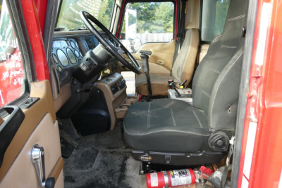 CV truck cab interior showing driver seat and steering wheel with beige door panels and dashboard; fire extinguisher under seat; gear shifter and pedals visible.