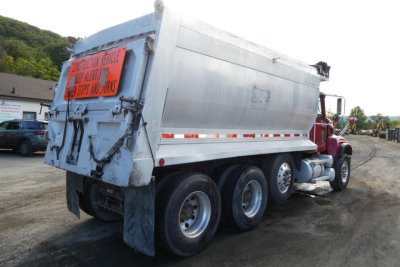 Dump truck rear and side view showing orange warning sign, hydraulic tailgate, and triple-axle rear. Exterior metal panels with reflective tape along the side.