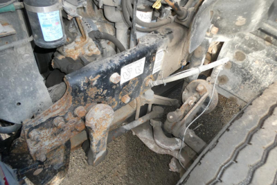 Mack CV Granite dump truck front suspension and steering linkage with rust and dirt, visible shock absorber, wheel, and hydraulic components.