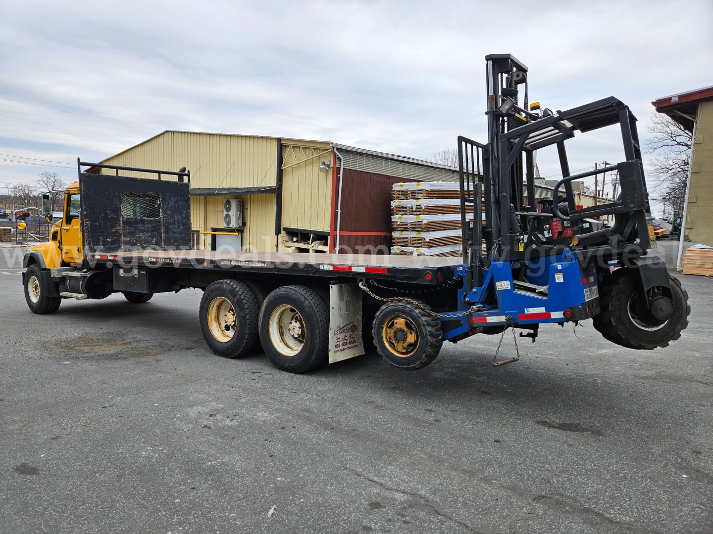 Flatbed truck with a large forklift mounted on a lowboy trailer; rear view shows three axles, yellow cab, black chassis, and forklift forks raised on blue frame.